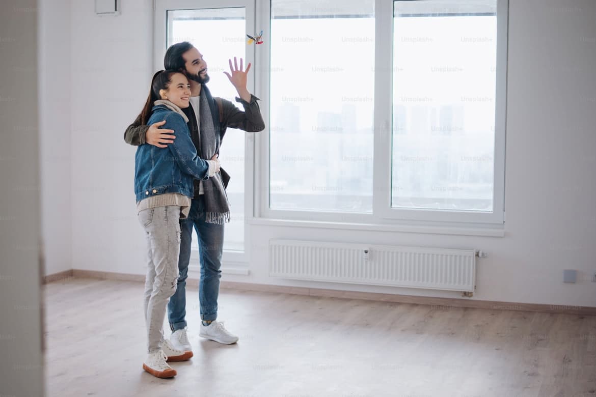 A happy young couple stands close together in a bright, empty house they just bought. They wear warm clothes and are smiling. The camera has caught them at the moment when someone is tossing them the keys.