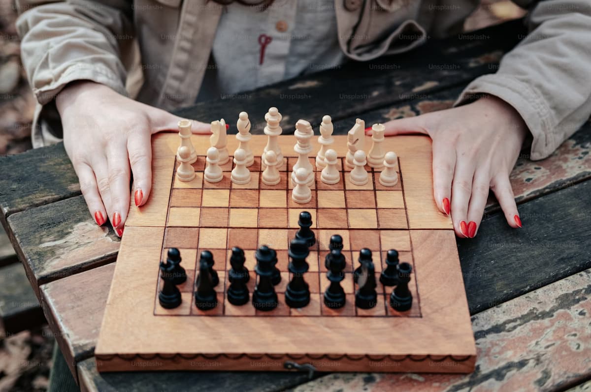 A woman’s hands rest on either side of a chess board. All of the game pieces are lined up, with a few of the pawns already moved.