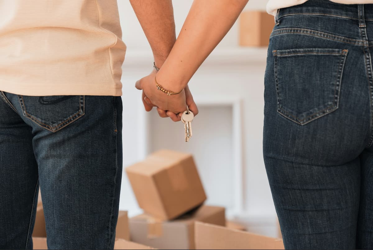 A couple holding hands, with a set of keys dangling from their fingers. They are both wearing jeans, and there are cardboard moving boxes in the background.