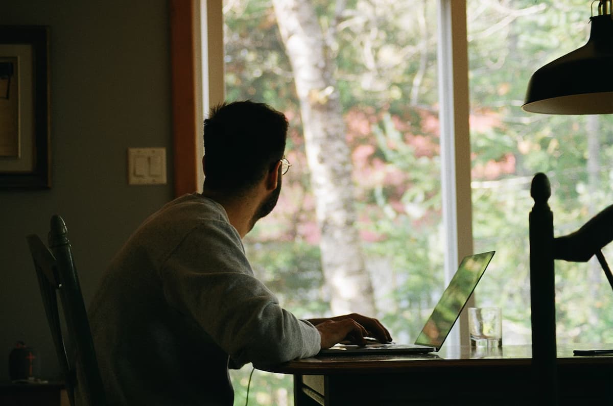 Man in a gray hoodie using a laptop looking out a window.
