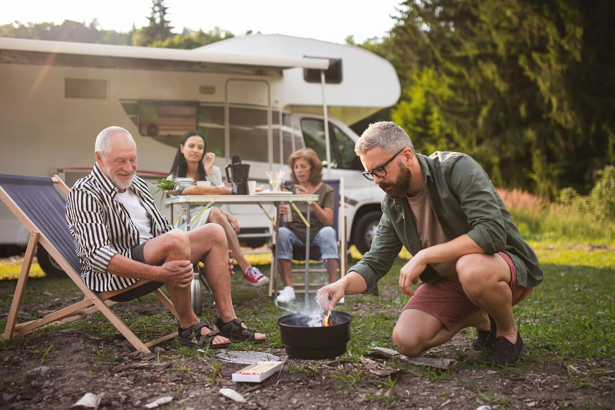 A family is camping with their RV, and a man is starting a small barbecue in the foreground.
