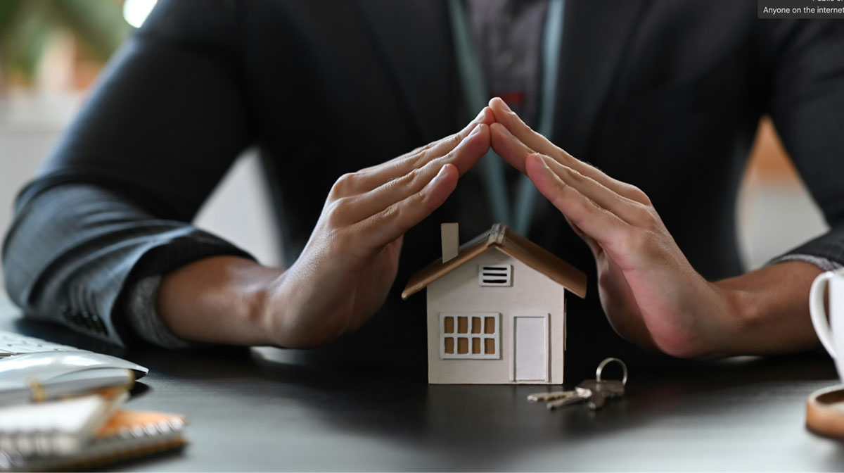 A person in a suit shields a model house with their hands.